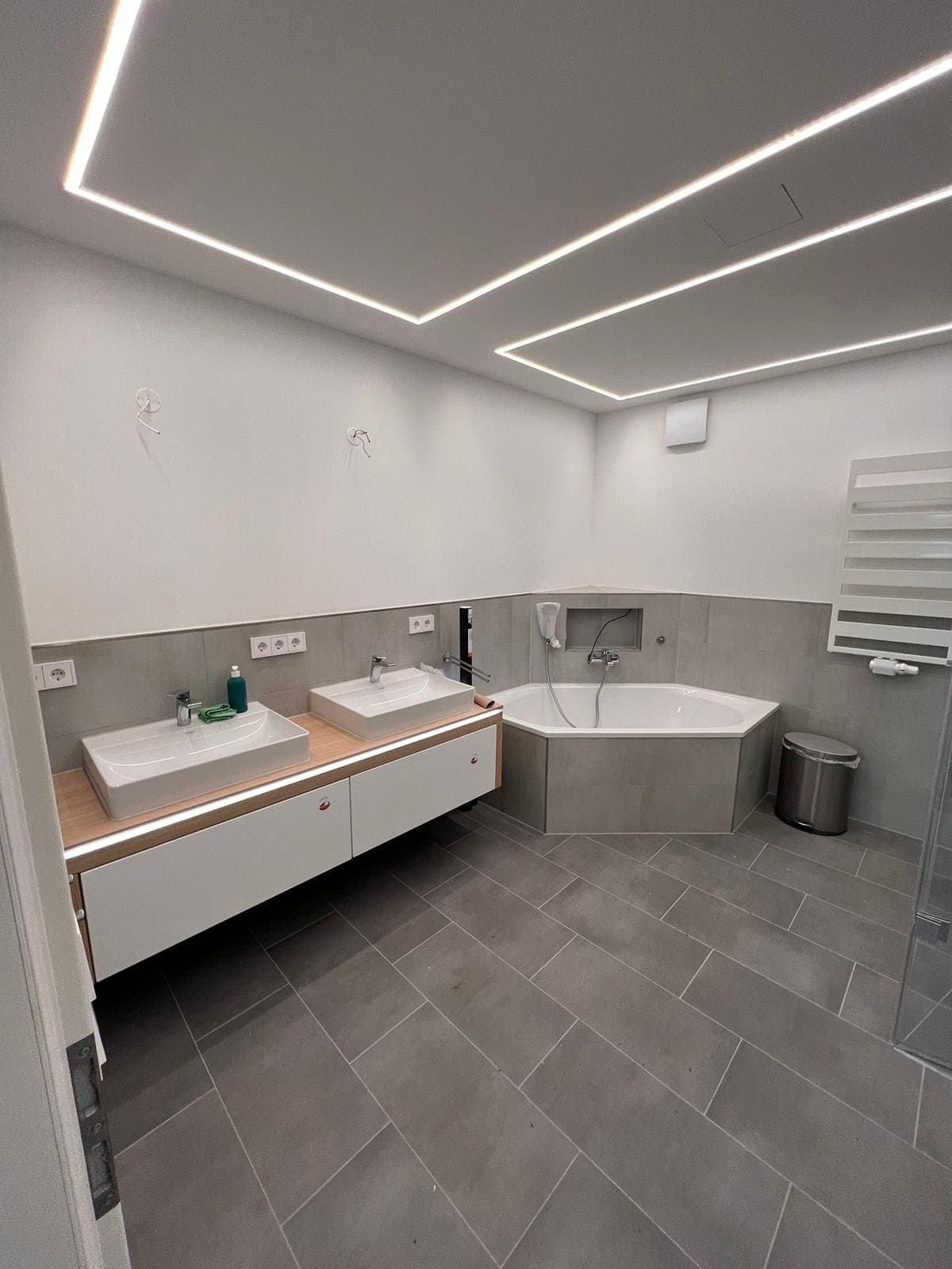 Modernes Badezimmer mit rechteckigen LED-Lichtlinien in der Decke, Doppelwaschtisch und Eckbadewanne